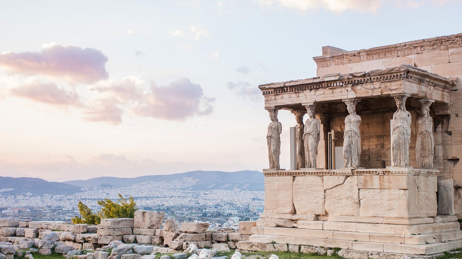 Ancient Greek temple ruins with stone columns shaped like human figures, set against a backdrop of a cityscape and rolling hills at sunset.