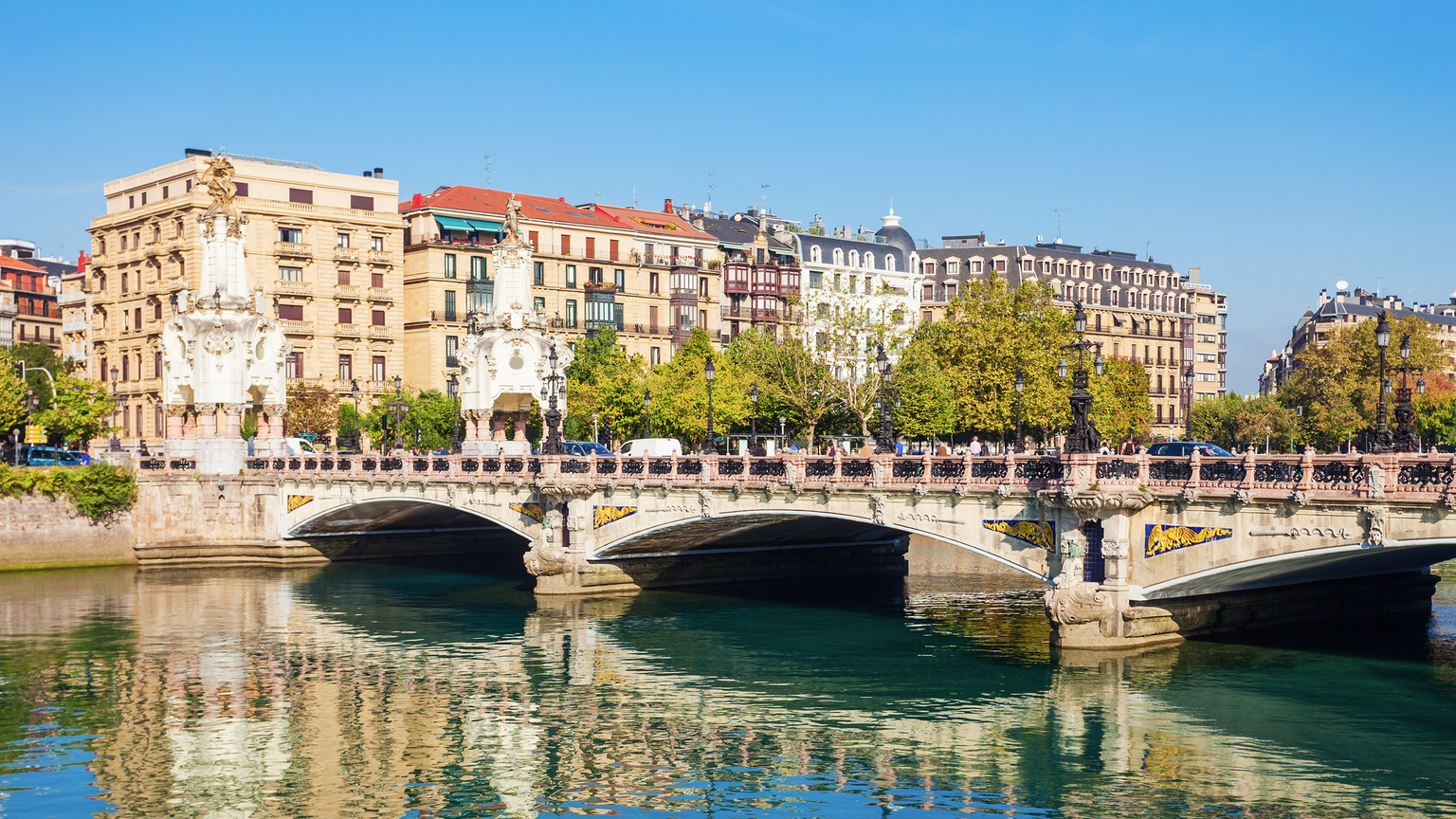 A scenic view of a historic stone bridge over a river with ornate buildings and trees under a clear blue sky in the background.