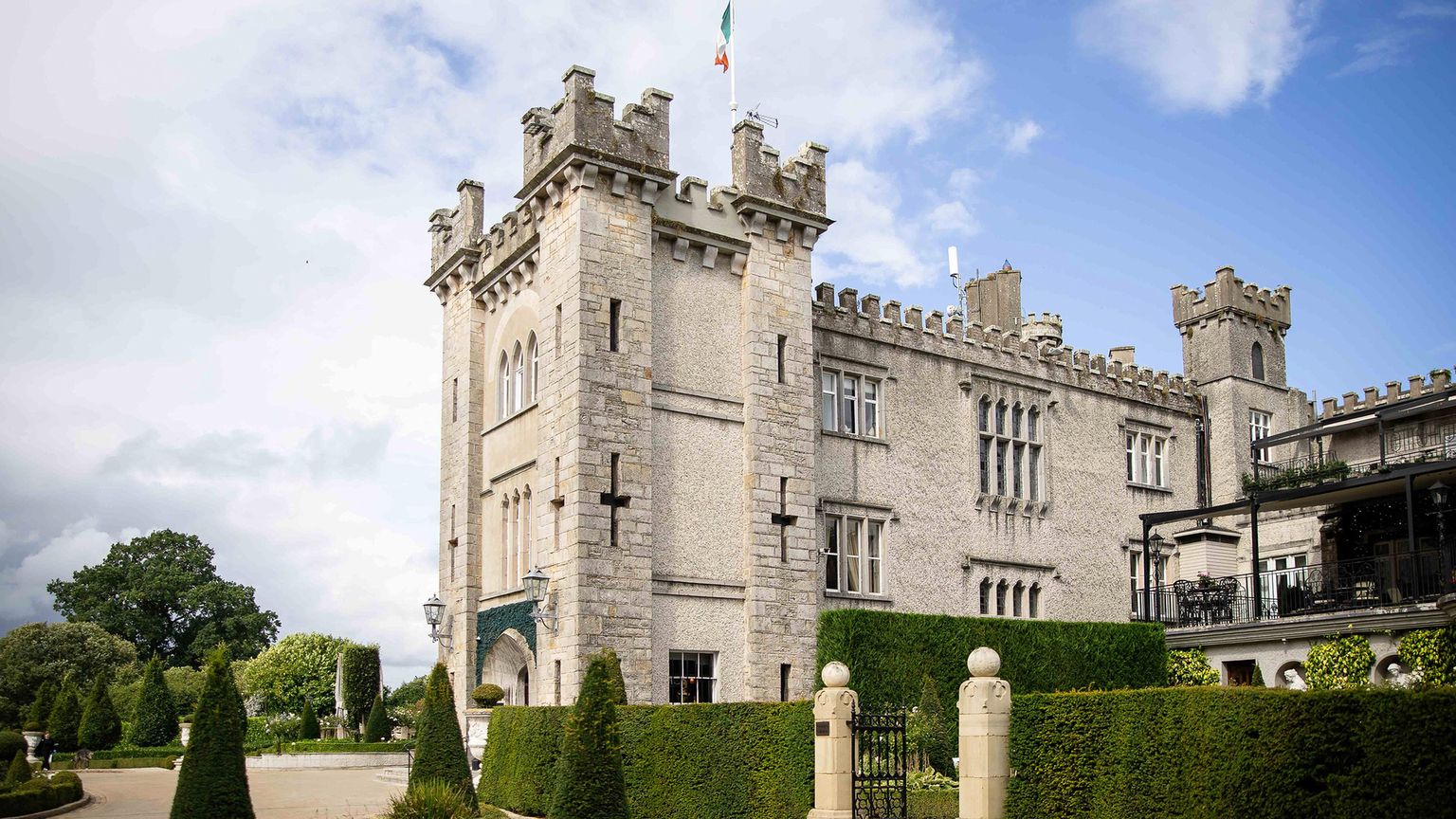 Stone castle with crenellated towers and an Irish flag, surrounded by manicured gardens under a blue sky.