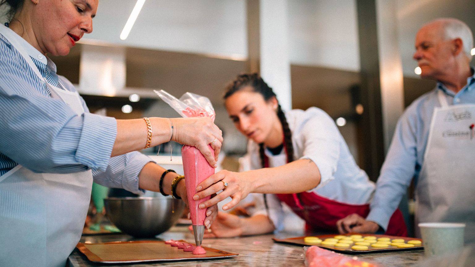 People in a kitchen, focused on piping pink batter onto trays, with bowls and other baking tools visible in the background.