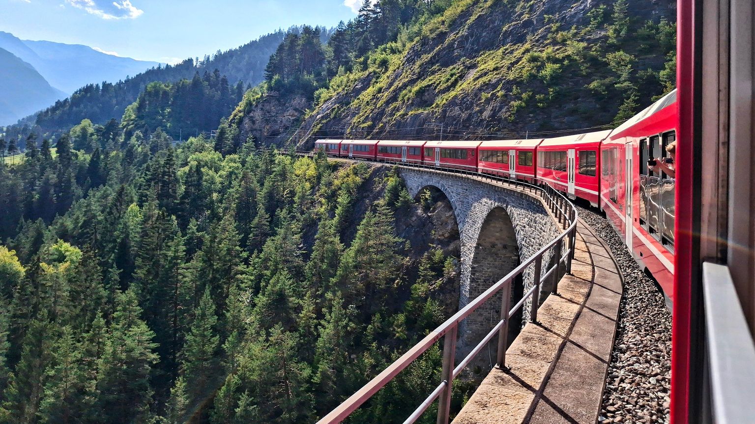 A red train travels along a curved stone bridge through a lush green forest, with mountains visible in the background under a clear sky.