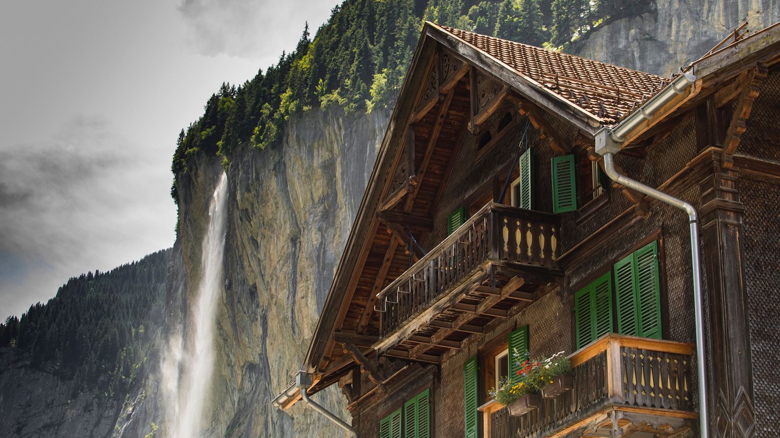 Rustic wooden house with green shutters in front of a dramatic cliff with a waterfall, surrounded by lush green forest.