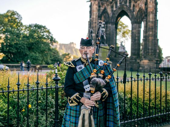 A bagpiper in traditional Scottish attire plays near an ornate stone arch, with lush greenery and a historic structure in the background.