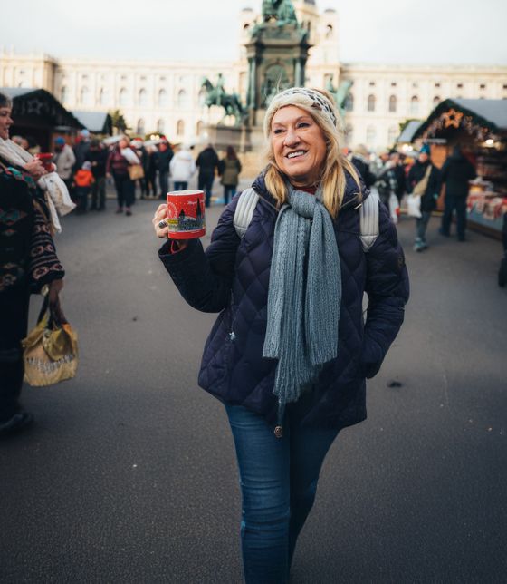 Smiling woman in winter attire holding a red mug, standing in a busy outdoor market with historic architecture in the background.