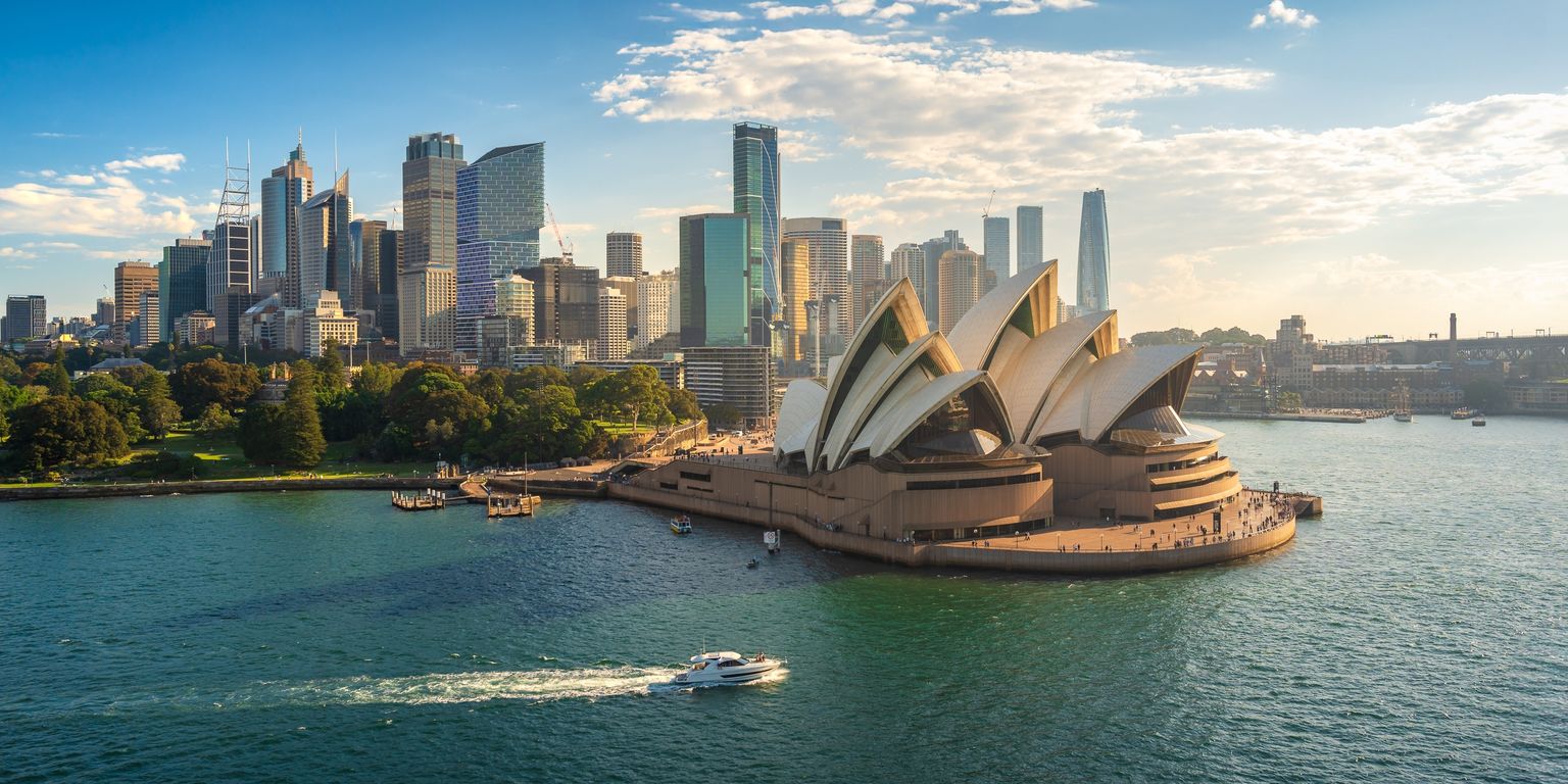 Sydney Opera House by the harbor with a boat on the water, city skyline in the background under a partly cloudy sky.