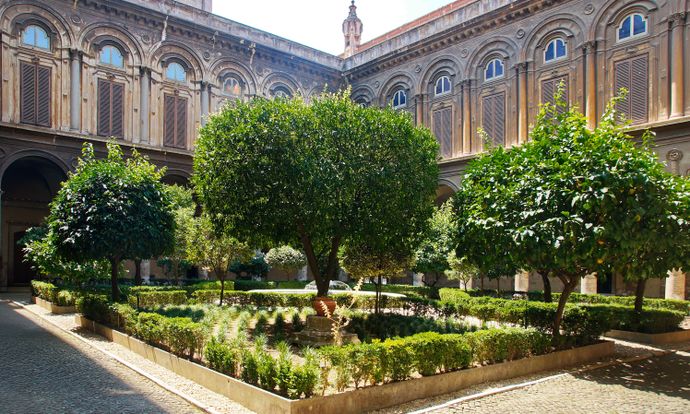 Lush courtyard with ornate architecture, featuring a central tree surrounded by trimmed hedges and smaller plants under a sunny sky.