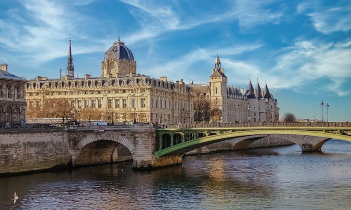 View of the historic Conciergerie and Pont au Change bridge over the Seine River in Paris, under a blue sky with scattered clouds.