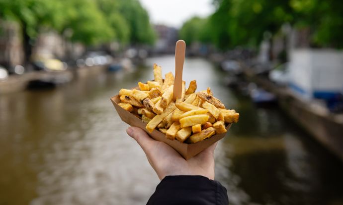 A hand holds a paper tray of fries with a wooden fork against a blurred background of a canal lined with trees.