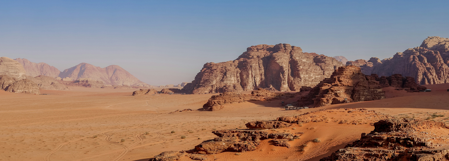 Desert landscape with large rock formations under a clear blue sky, featuring orange sand dunes and rugged cliffs in the distance.