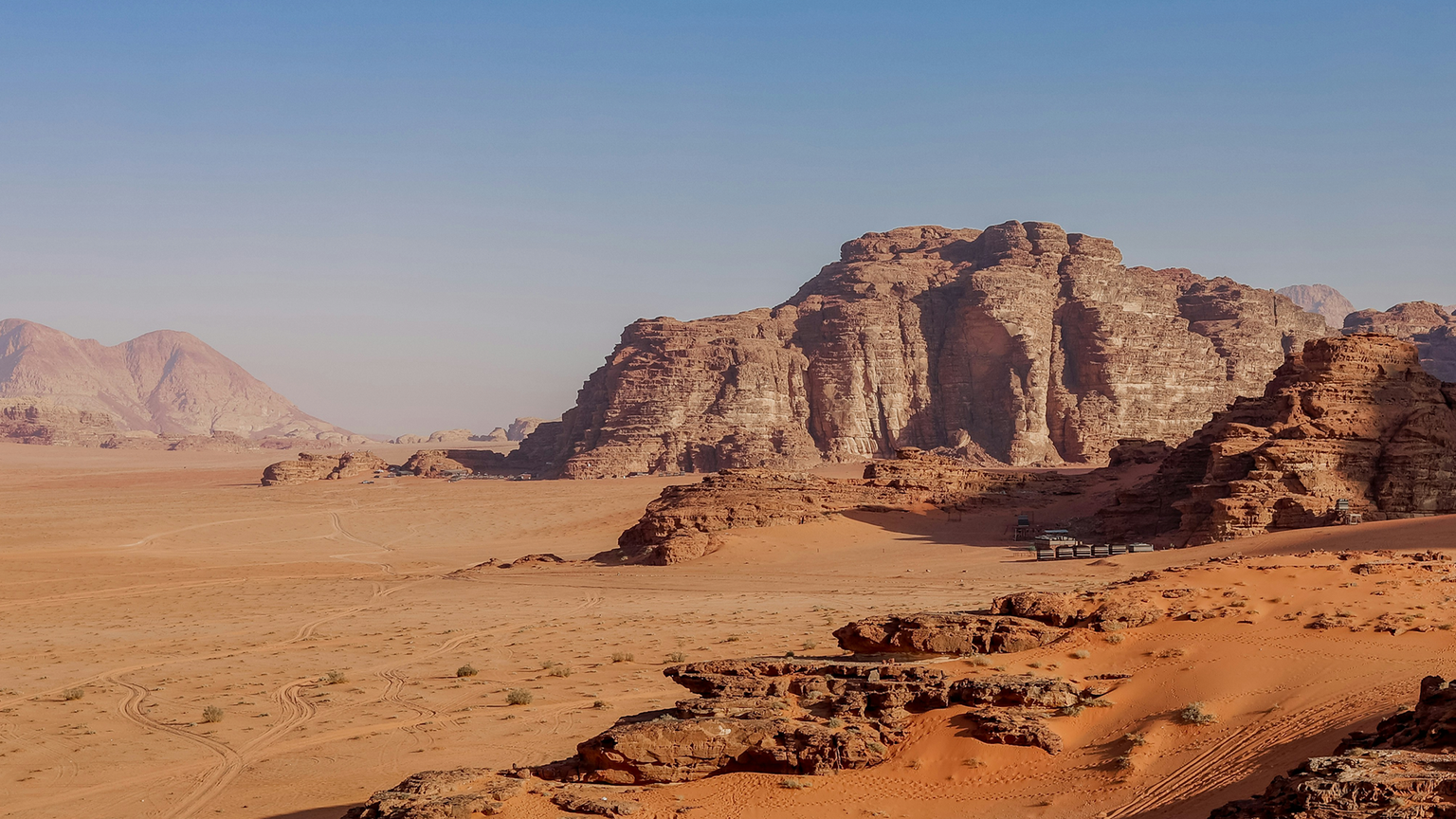 Desert landscape with large rock formations under a clear blue sky, featuring orange sand dunes and rugged cliffs in the distance.