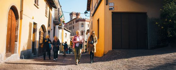 Four travelers walk along a sunlit cobblestone street between colorful historic buildings, with a medieval castle visible in the background.