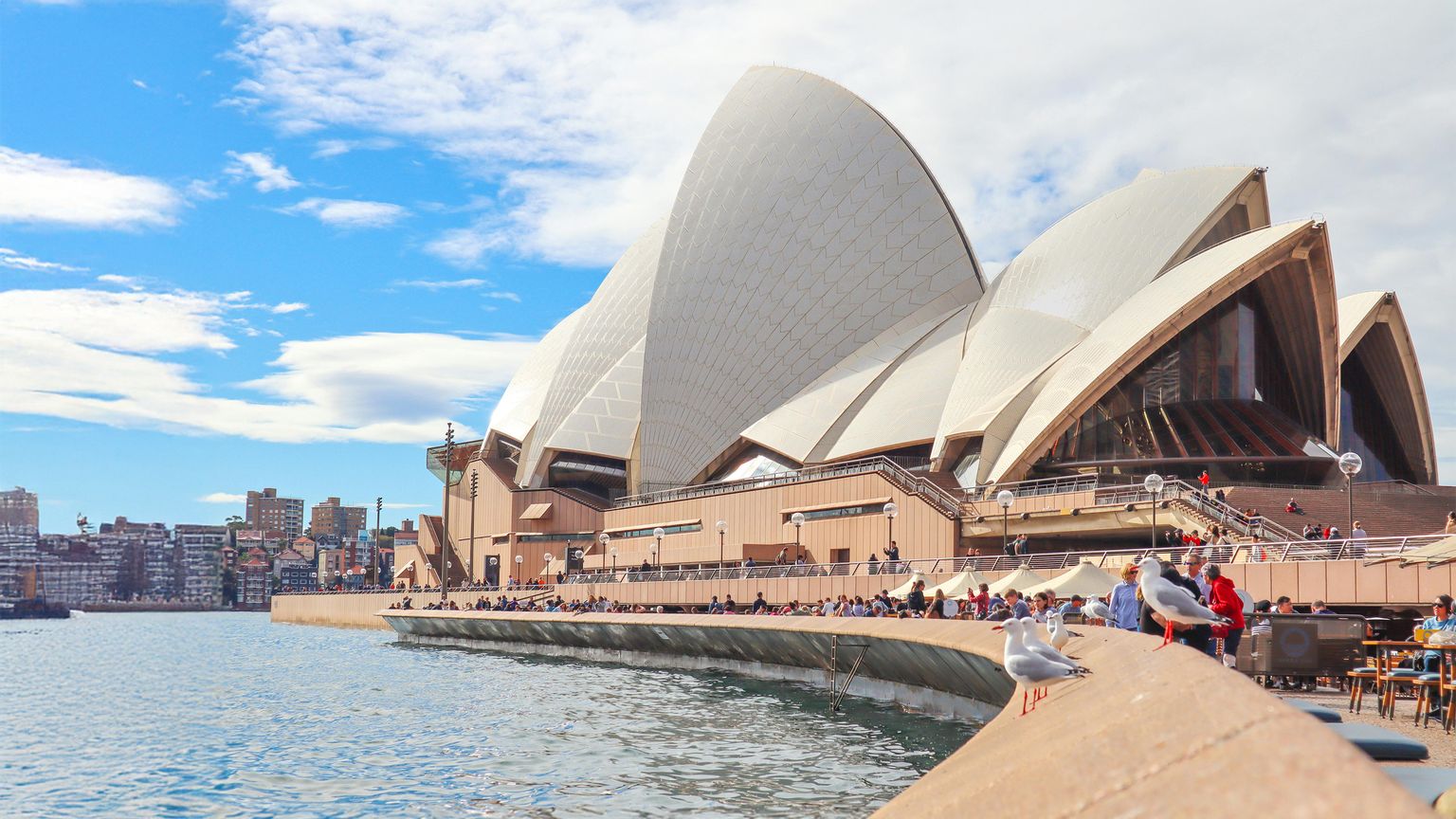Sydney Opera House by the waterfront, with people and seagulls on the promenade under a partly cloudy blue sky.