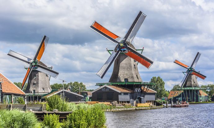 Three traditional windmills with orange sails stand by a river under a cloudy sky, surrounded by greenery.