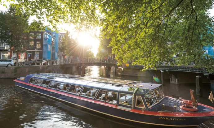 A canal boat with passengers navigates an Amsterdam waterway, surrounded by historic buildings and lush trees, under the warm glow of the setting sun.