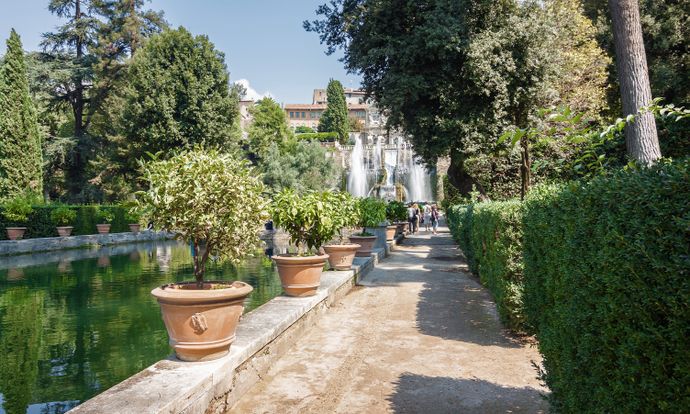 Pathway lined with potted plants beside a pond, leading to a cascading fountain with lush trees and a building in the background.