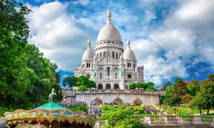 Sacré-Cœur Basilica in Paris, framed by blue sky and clouds, with a carousel in the foreground and lush greenery surrounding it.