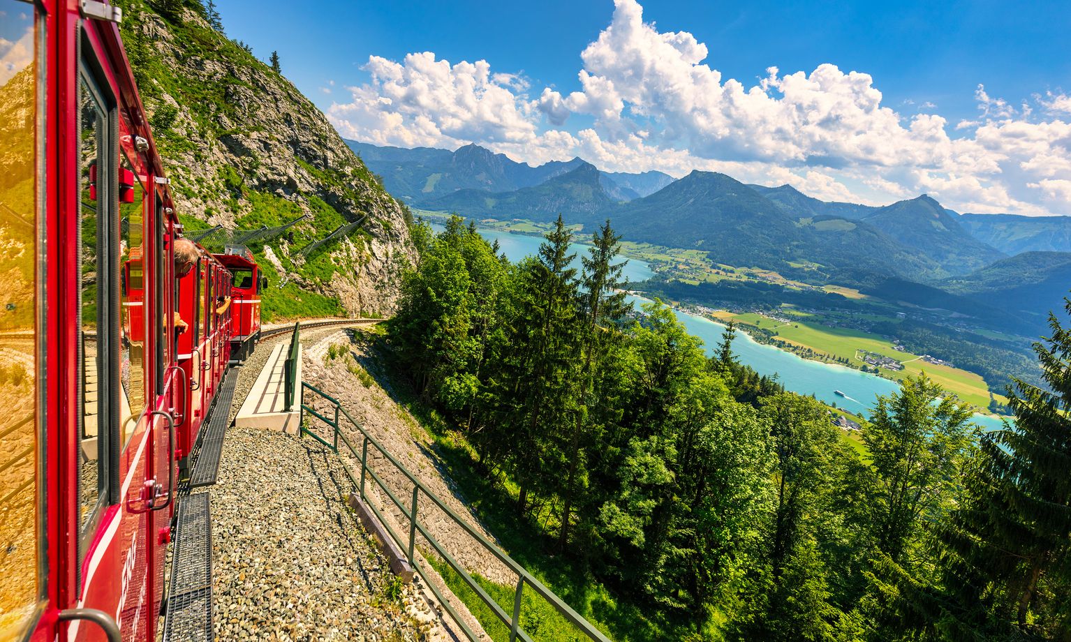 A red train travels along a mountainside, overlooking a scenic landscape of lush greenery, a blue lake, and distant mountains under a cloudy sky.