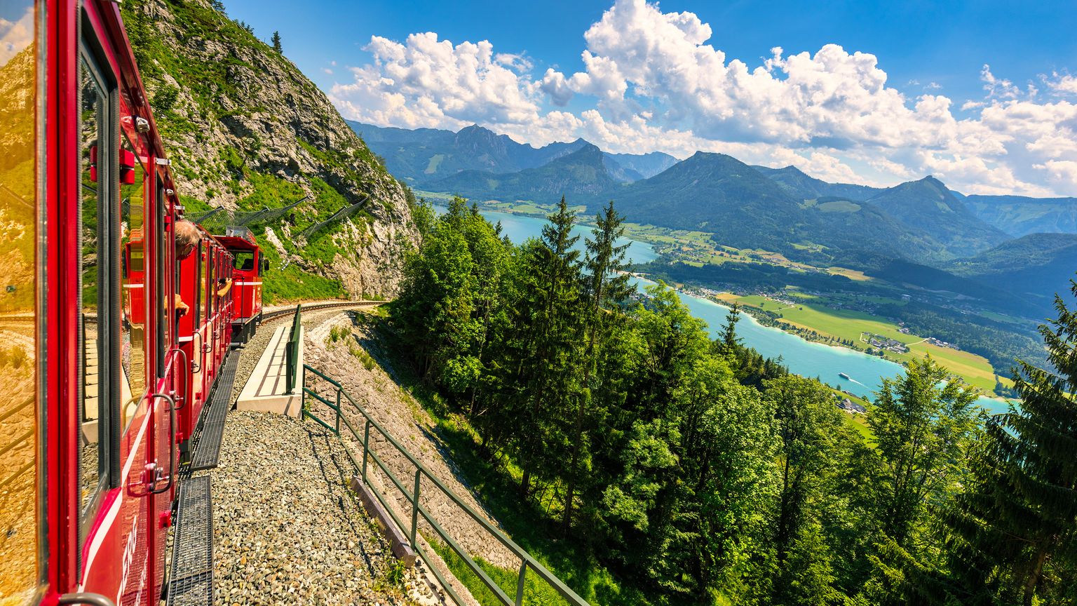 A red train travels along a mountainside, overlooking a scenic landscape of lush greenery, a blue lake, and distant mountains under a cloudy sky.