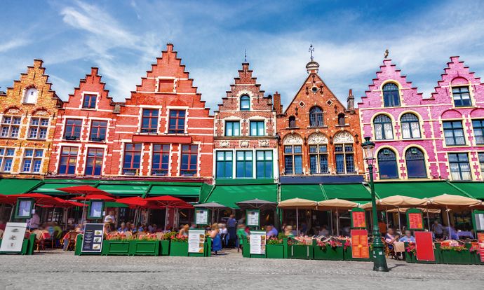Colorful gabled buildings with outdoor cafes under red and green canopies line a cobblestone street against a blue sky.