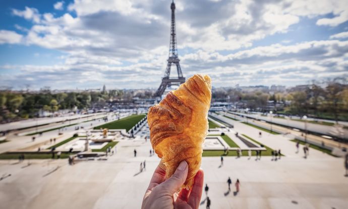 A hand holds a croissant in front of a blurred Eiffel Tower, with a view of park grounds and a partly cloudy sky.