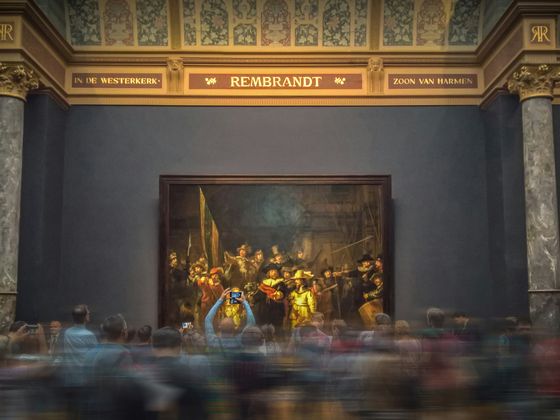 People gathered in front of Rembrandt's "The Night Watch" at a museum, with blurred motion indicating movement within the crowd.