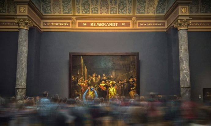 People gathered in front of Rembrandt’s “The Night Watch” at a museum, with blurred motion indicating movement within the crowd.