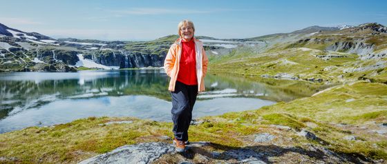A person in a bright jacket stands on a rock, smiling near a tranquil lake with snow-capped mountains under a clear blue sky.