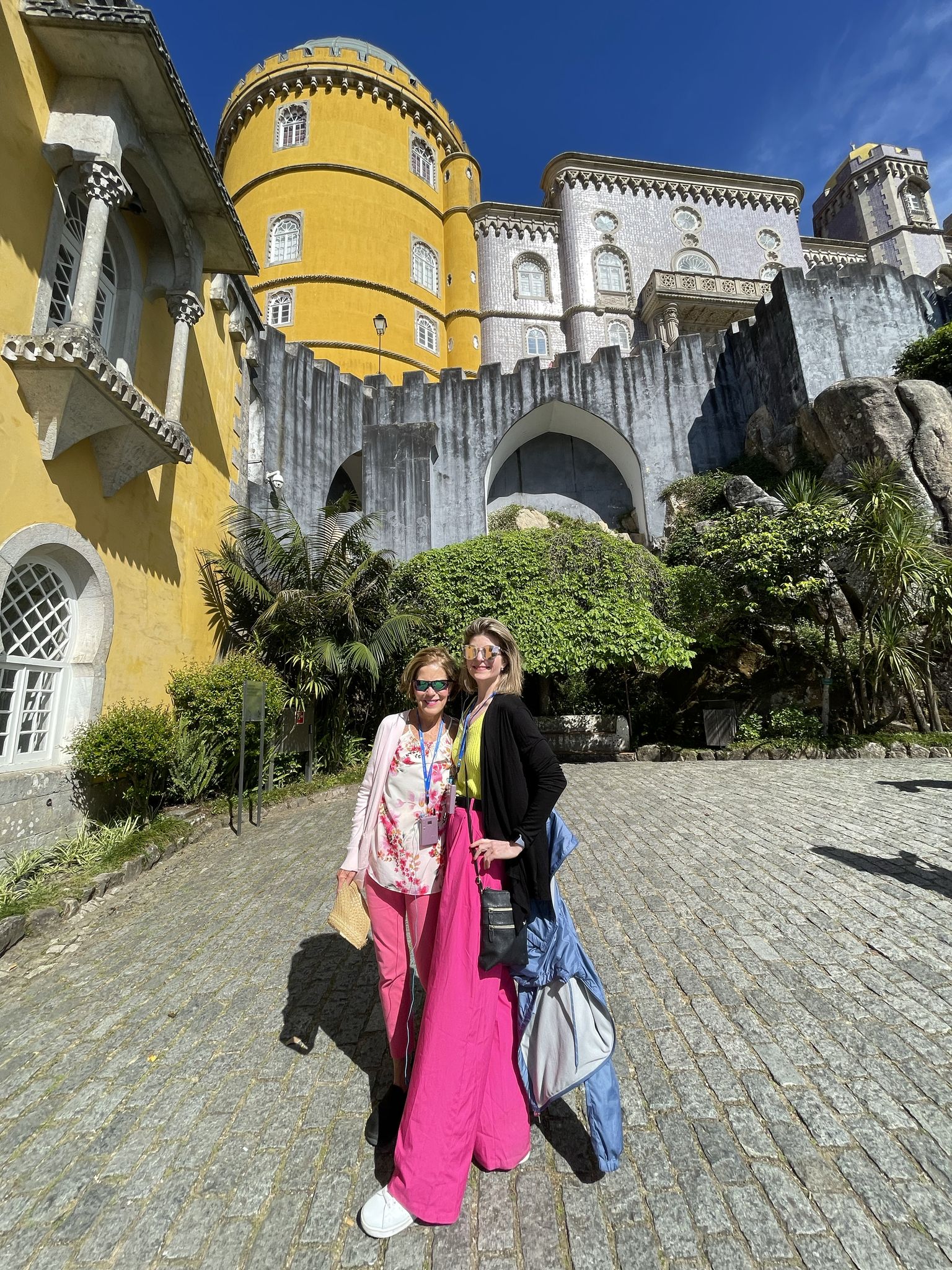 Two people smiling and posing in front of a colorful, historic castle with round towers and decorative arches, under a clear blue sky.