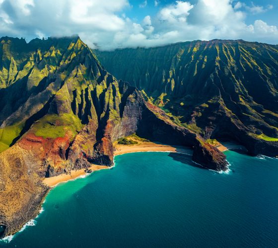 Aerial view of lush, jagged cliffs with vibrant green and red hues along a turquoise shoreline, under a partly cloudy sky.