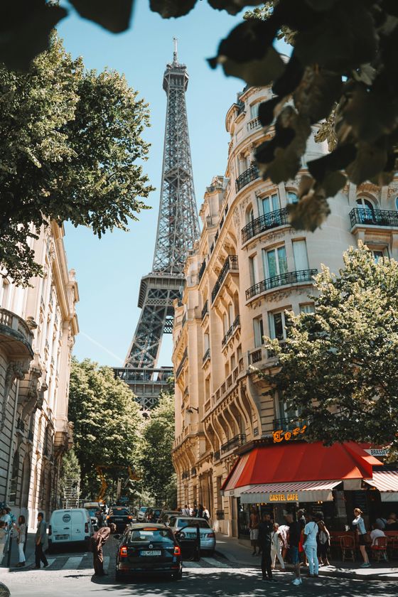 Street view with Parisian architecture, a red-awning café, cars, and people, with the Eiffel Tower rising prominently in the background.