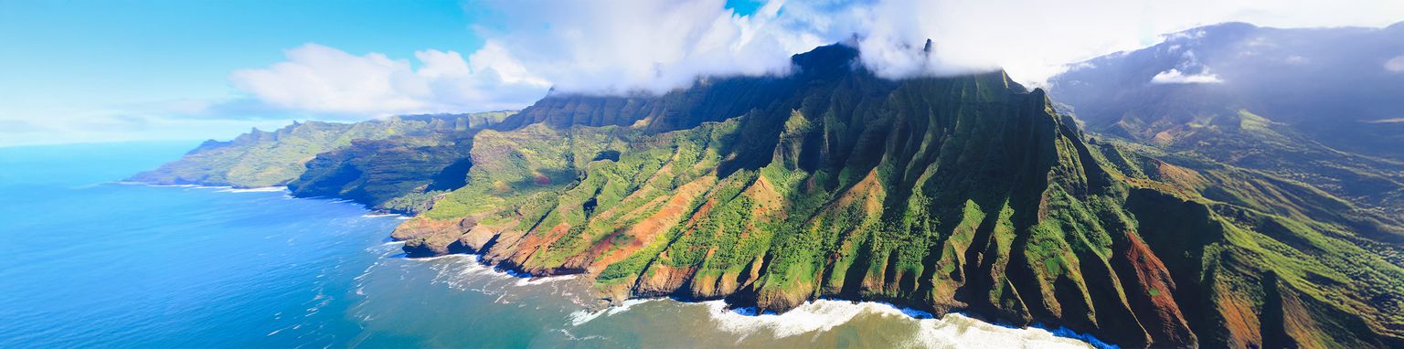Aerial view of Na Pali Coast in Hawaii with dramatic cliffs, lush greenery, and blue ocean waves under a partly cloudy sky.
