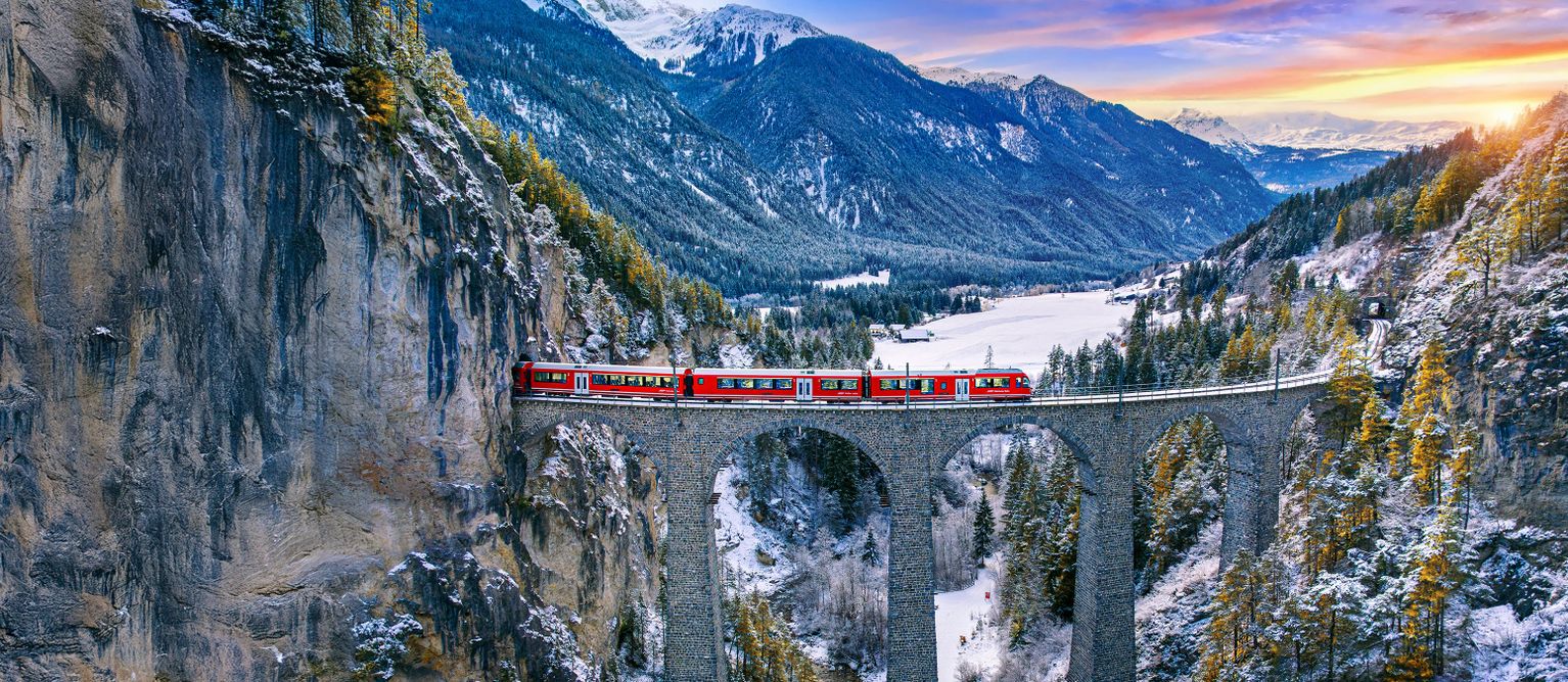 A red train crosses a stone viaduct in a snowy mountain landscape at sunset, surrounded by steep cliffs and pine trees.