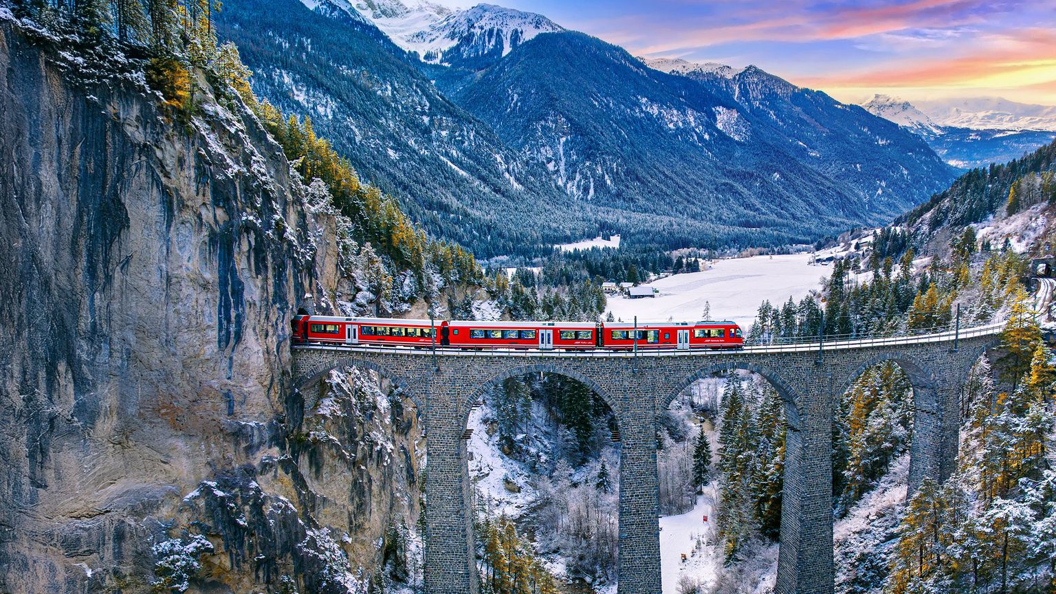 A red train crosses a stone viaduct in a snowy mountain landscape at sunset, surrounded by steep cliffs and pine trees.