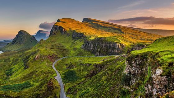 Scenic view of Quiraing, Isle of Skye, with winding road, green hills, and rocky cliffs at sunset under a colorful sky.