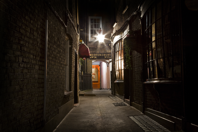 A dimly lit narrow alley with brick walls, leading to a warmly lit entrance under a sign that reads "Bedfordbury Wine Bar."