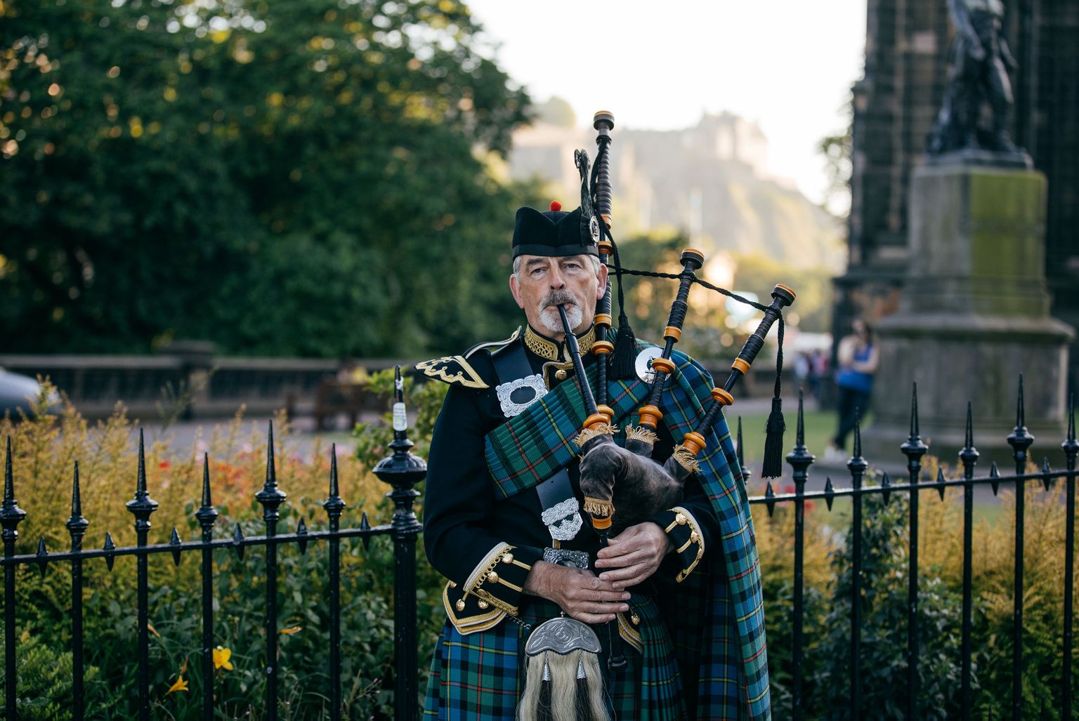 A man in traditional Scottish attire plays a bagpipe in a park, surrounded by a fence and greenery, with a historic building in the background.