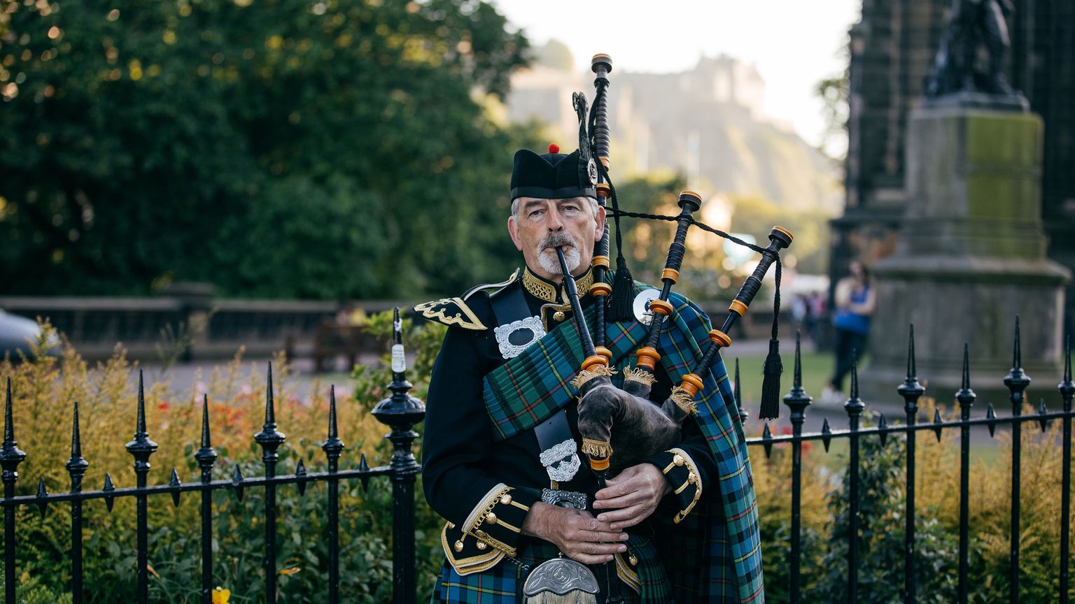 A man in traditional Scottish attire plays a bagpipe in a park, surrounded by a fence and greenery, with a historic building in the background.