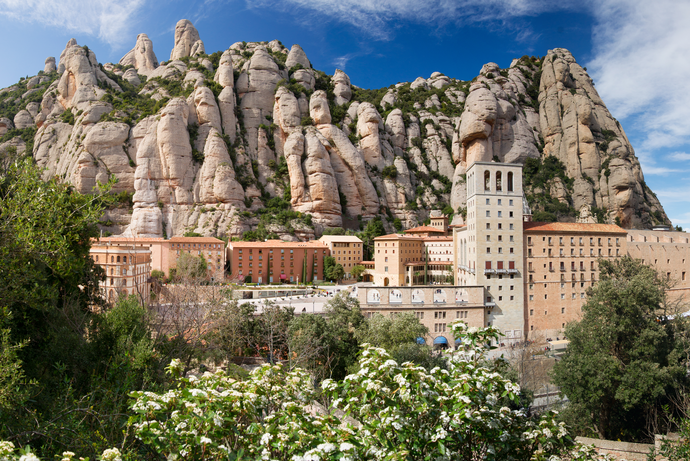 Monastery nestled against towering, jagged rock formations under a blue sky, with green foliage in the foreground.