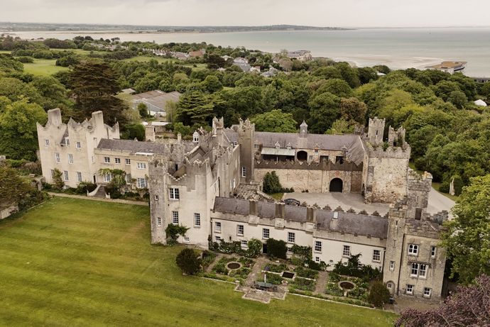 Aerial view of a historic castle with a courtyard surrounded by lush greenery, overlooking the sea in the background.