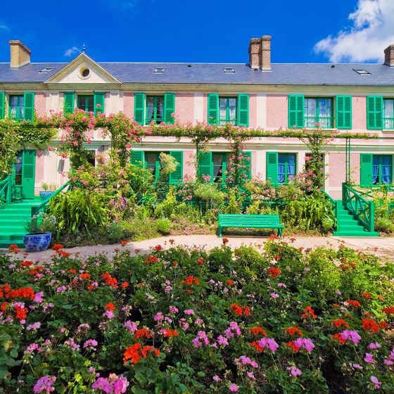 A vibrant garden with red and pink flowers in front of a large house with green shutters and a floral facade under a blue sky.