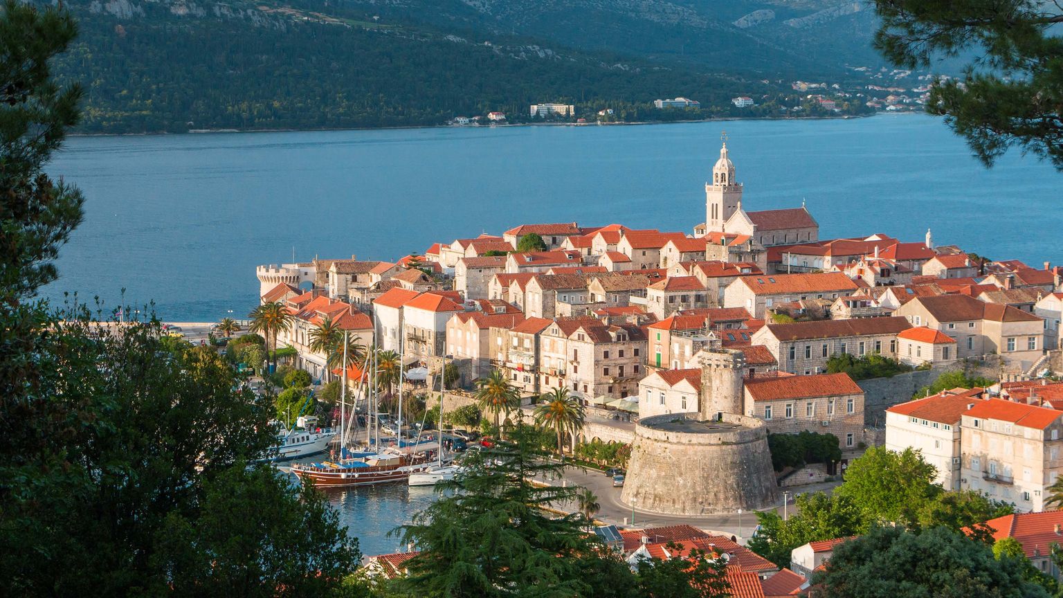 Scenic view of a coastal town with red-roofed buildings, a prominent church, and a harbor with boats, surrounded by mountains and blue water.
