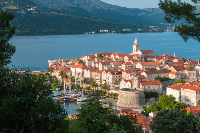 Scenic view of a coastal town with red-roofed buildings, a prominent church, and a harbor with boats, surrounded by mountains and blue water.