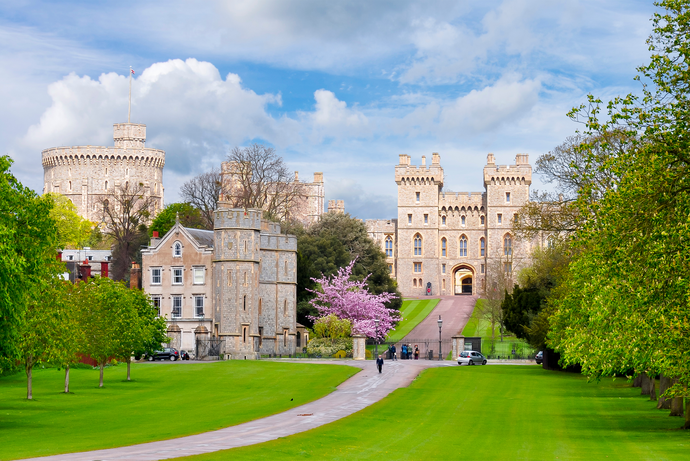Path leading to Windsor Castle, surrounded by lush green lawns and trees, with a blooming pink tree in the foreground under a partly cloudy sky.
