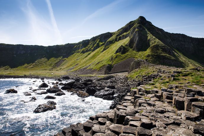 Scenic view of Giant's Causeway with hexagonal basalt columns, rocky shoreline, and a grassy hill under a clear blue sky.