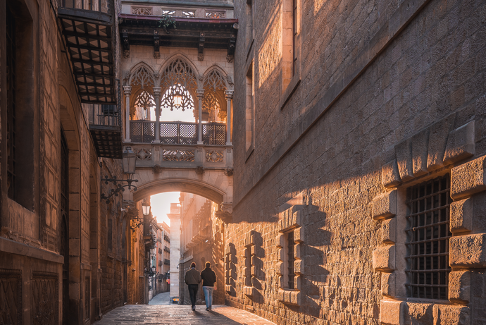 A narrow, sunlit medieval street with Gothic architecture and two people walking beneath an ornate stone bridge.