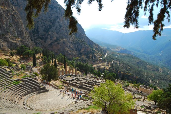 Ancient Greek amphitheater with stone ruins, surrounded by mountains and trees under a clear sky.