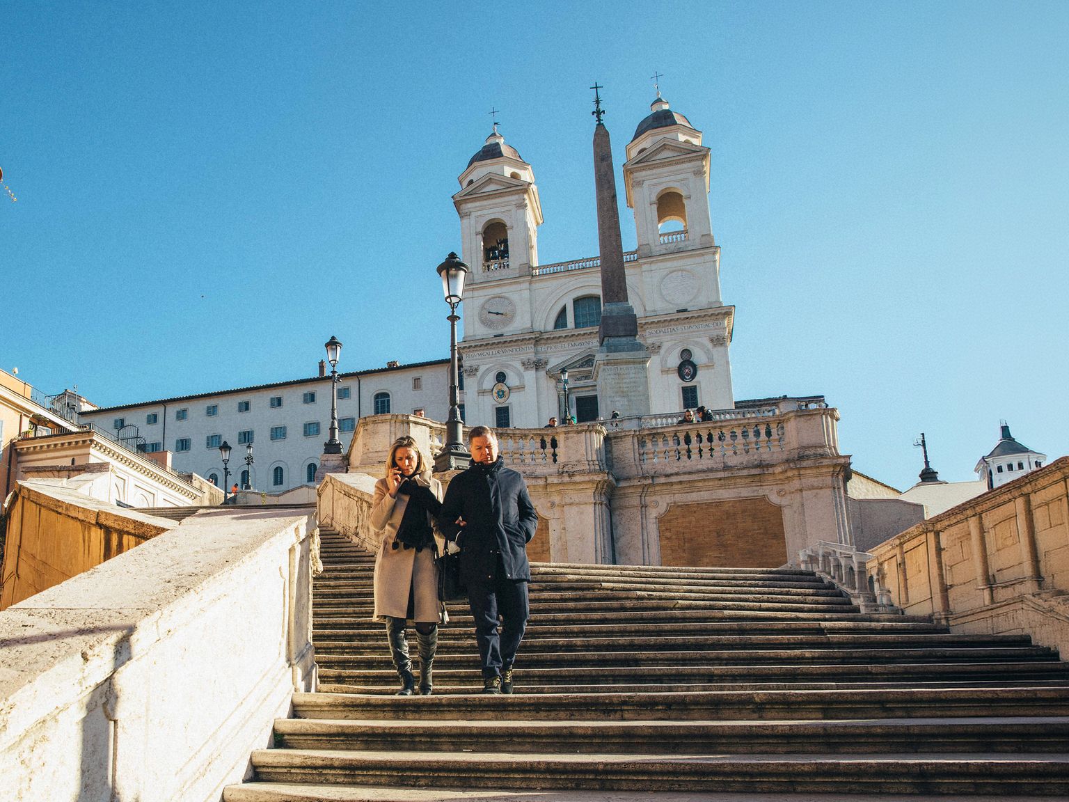 A couple walks down the Spanish Steps in Rome on a sunny day, with the Trinità dei Monti church in the background.