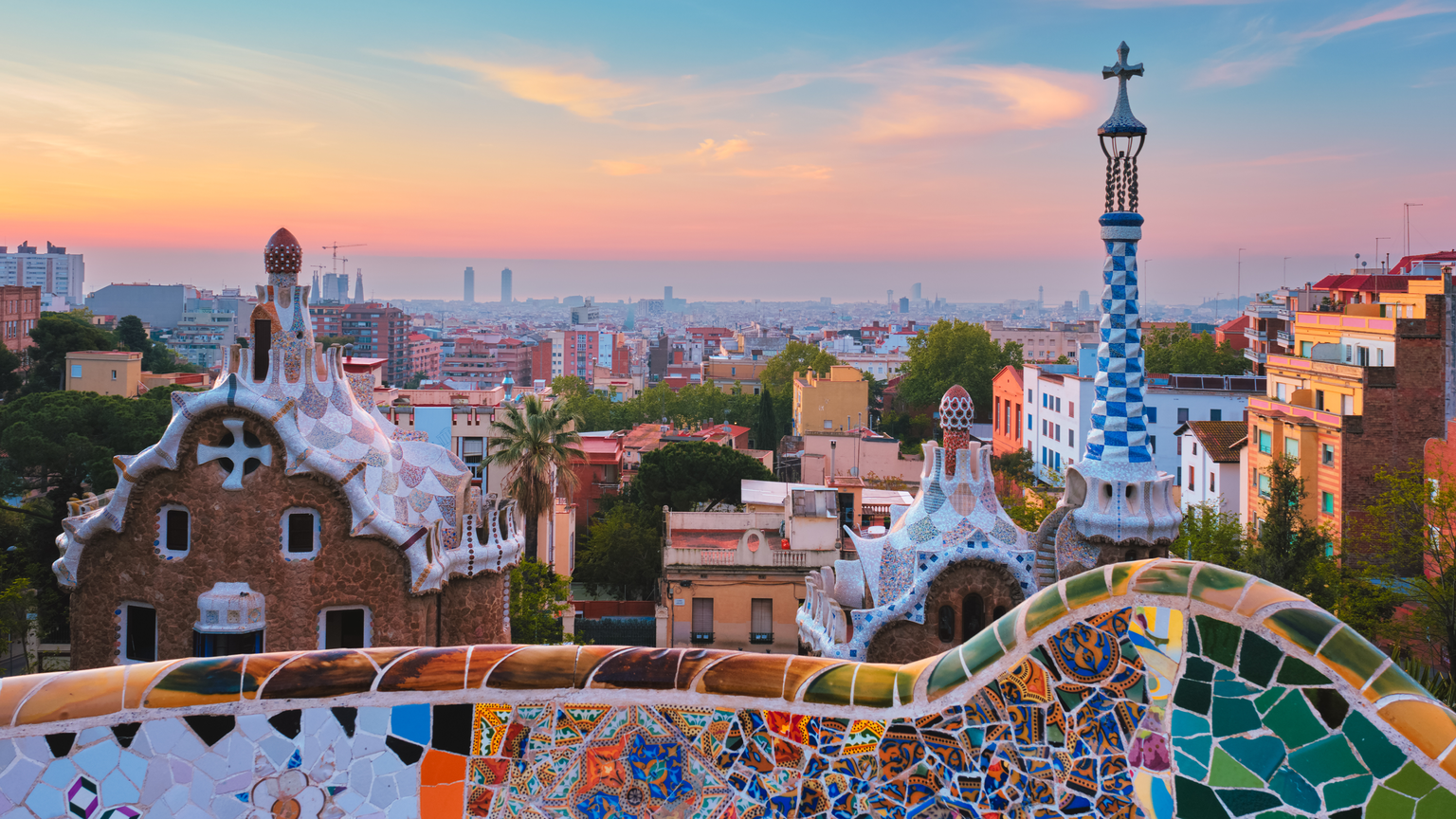 Colorful mosaic and unique architecture in Park Güell, Barcelona, with a cityscape and vibrant sunset sky in the background.