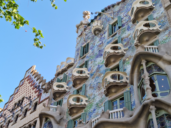 A colorful, ornate building facade with organic, curved balconies and mosaic tiles under a blue sky with some tree leaves visible.
