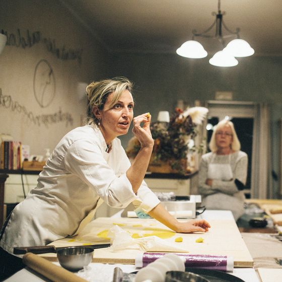A woman in a chef's coat teaches pasta-making to three women in aprons in a warmly lit kitchen setting.